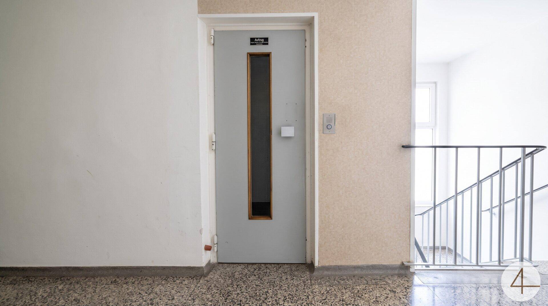 Aufzugtür in einem hellen Treppenhaus mit Zugang zu den oberen Etagen.