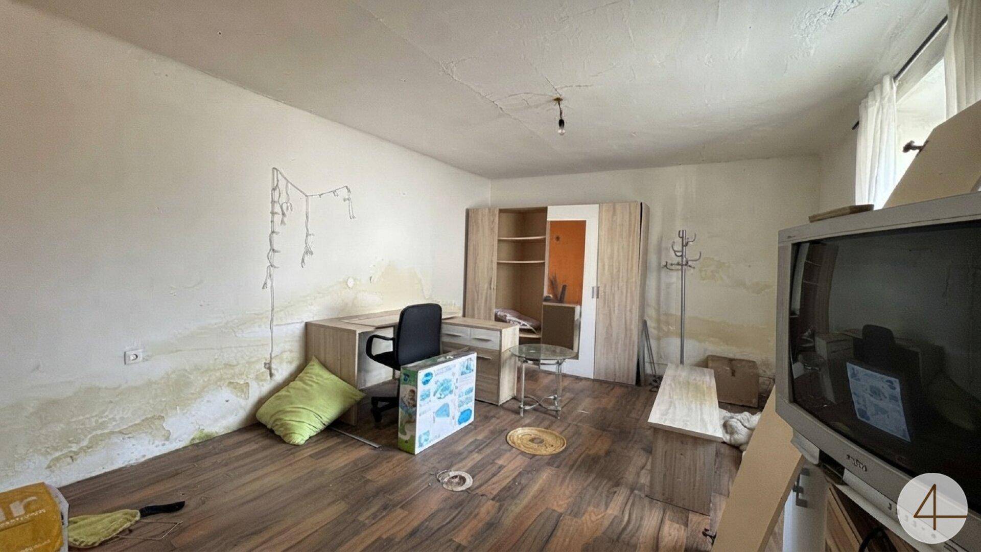 Zimmer mit Holzboden, Schreibtisch und Schrank, die Wände zeigen deutliche Feuchtigkeitsspuren.