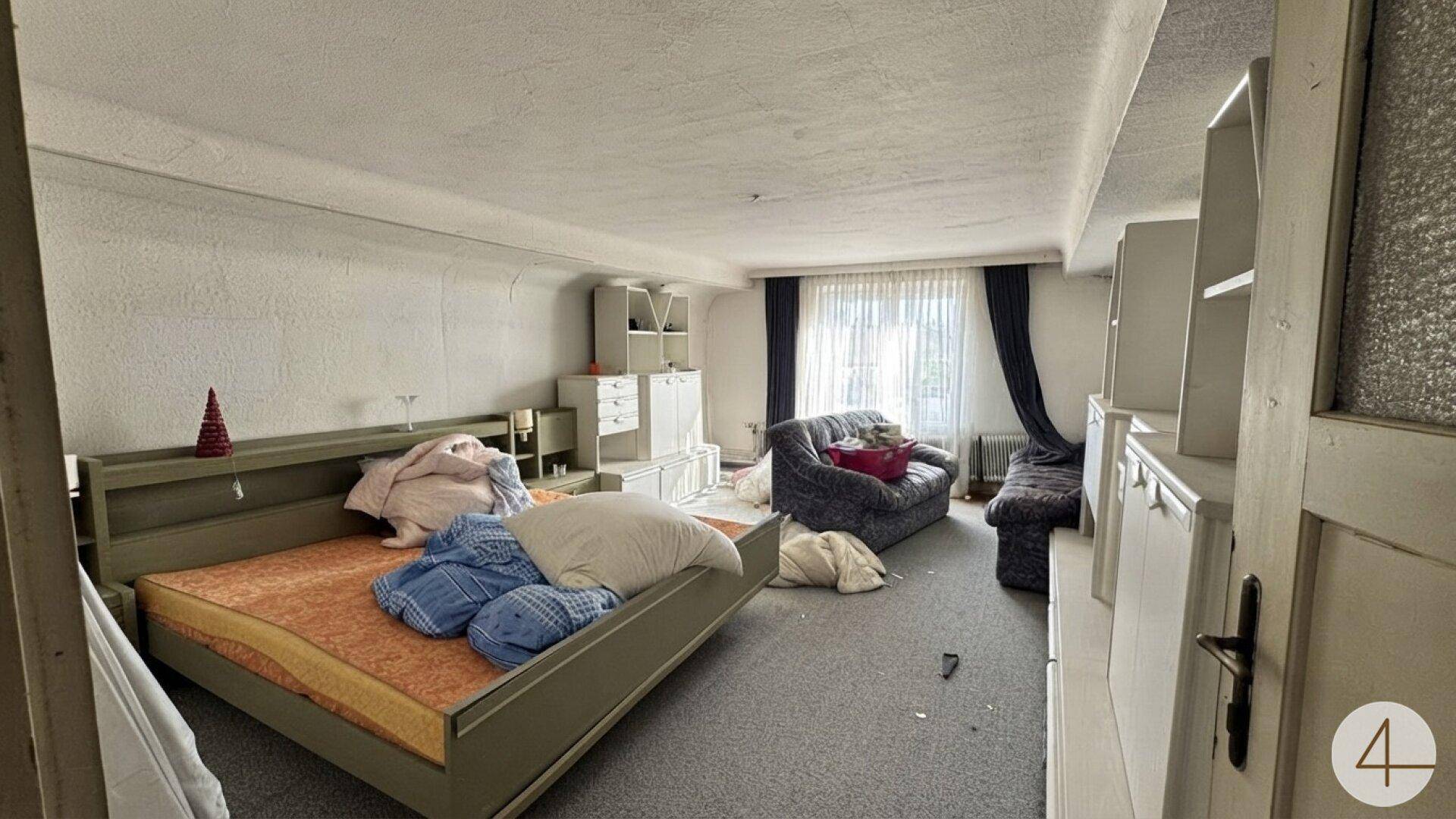 Schlafzimmer mit Teppichboden, Bett und Sofa, wirkt unaufgeräumt und renovierungsbedürftig.