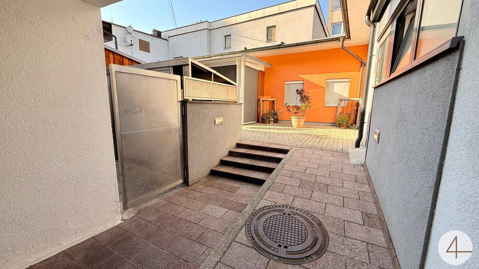 Gepflasterter Außenbereich zwischen Gebäuden mit Zugang zu einem Eingang und einer Treppe.