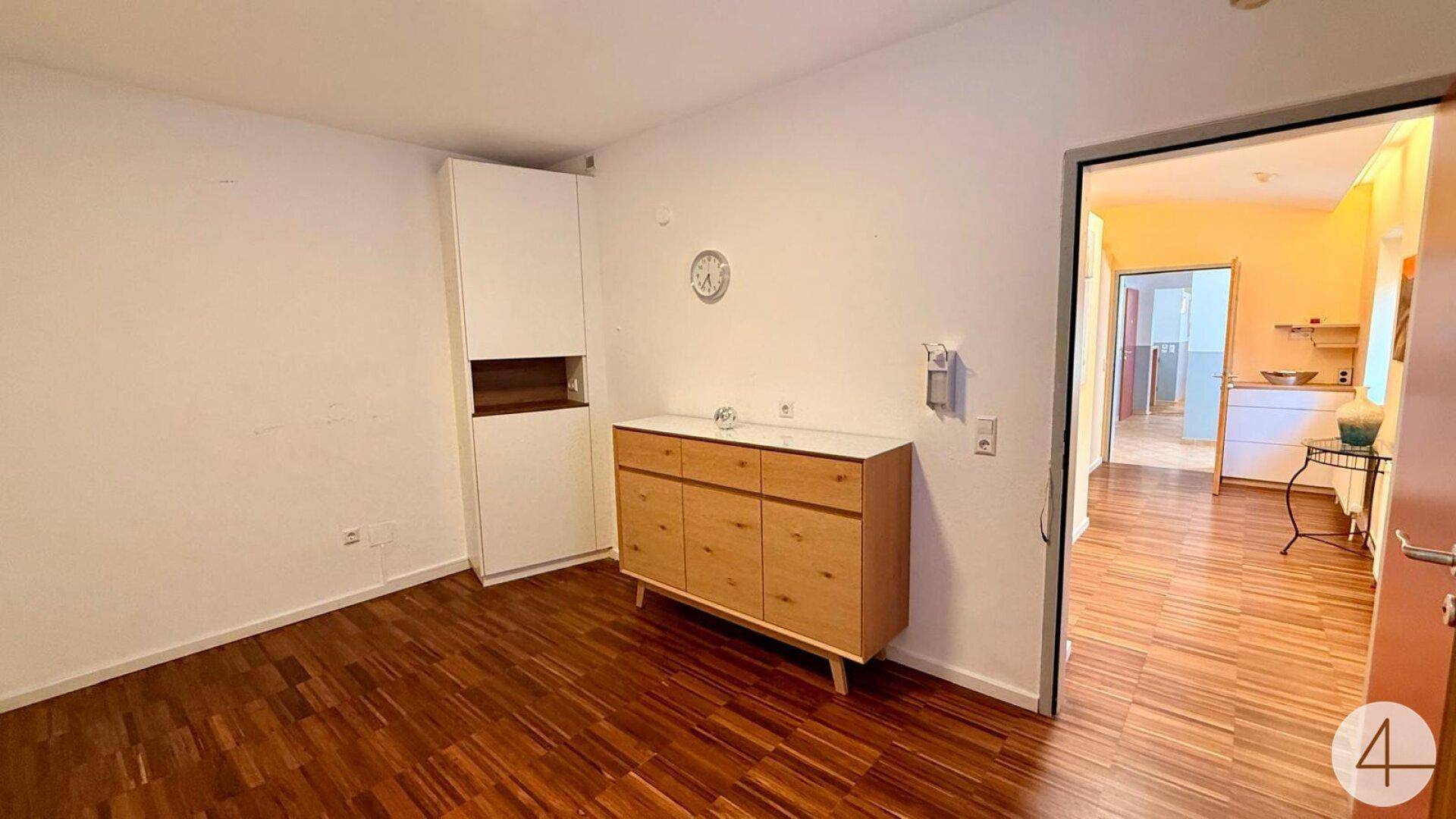 Heller Raum mit Holzfußboden, einem weißen Schrank und einer Holzkommode, führt in einen weiteren Raum.