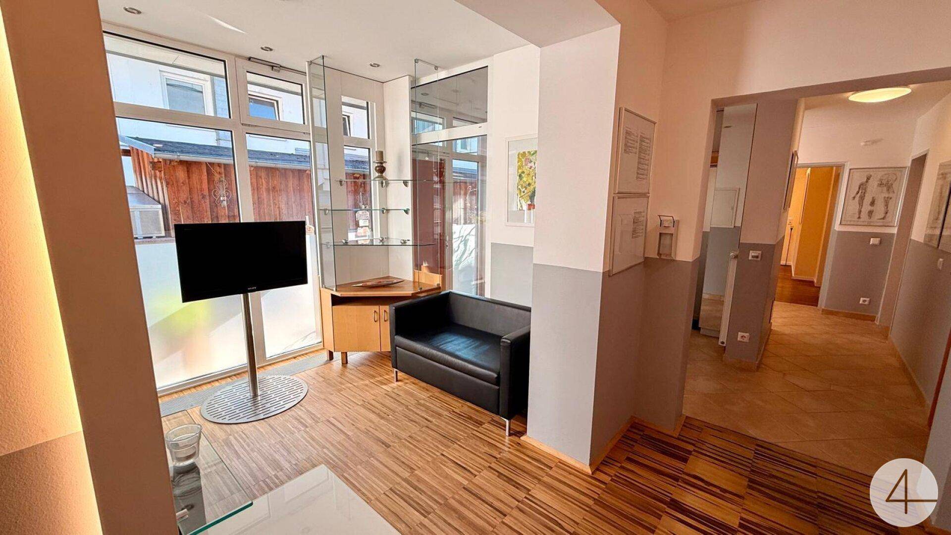 Wohnbereich mit Holzfußboden, schwarzem Sofa und großen Fenstern, die den Blick nach draußen freigeben.