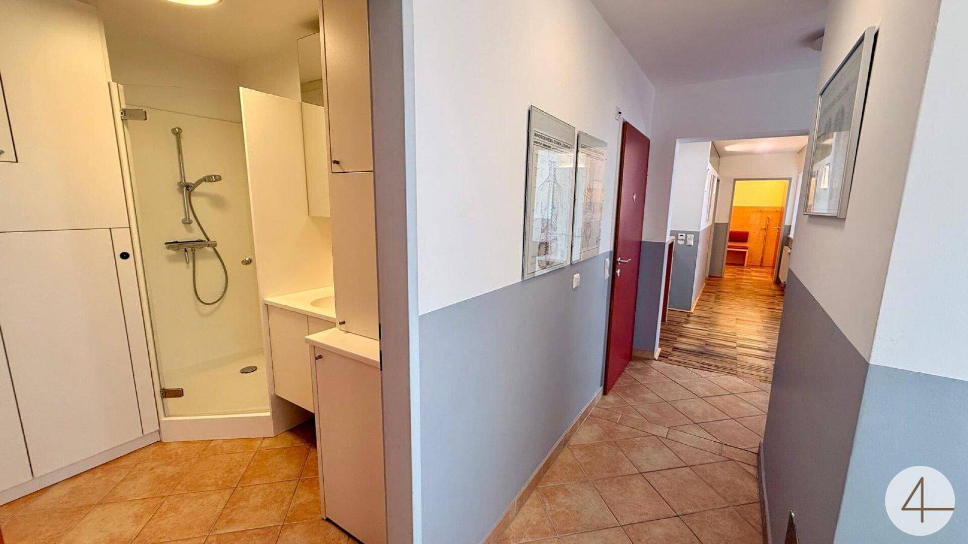 Flur mit Fliesenboden und Blick in einen Badezimmerbereich mit Dusche und weißen Schränken.