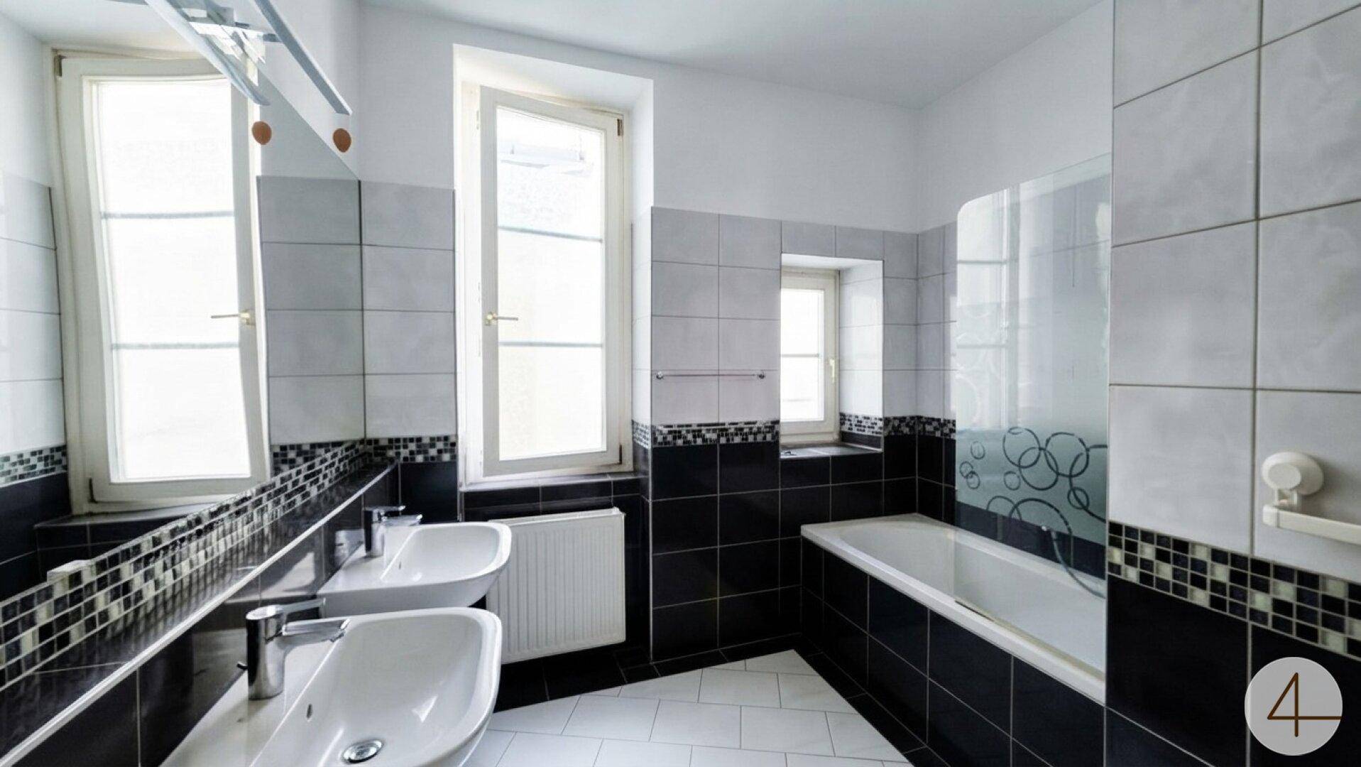 Modernes Badezimmer mit schwarz-weißen Fliesen, Doppelwaschbecken und Badewanne unter den Fenstern.