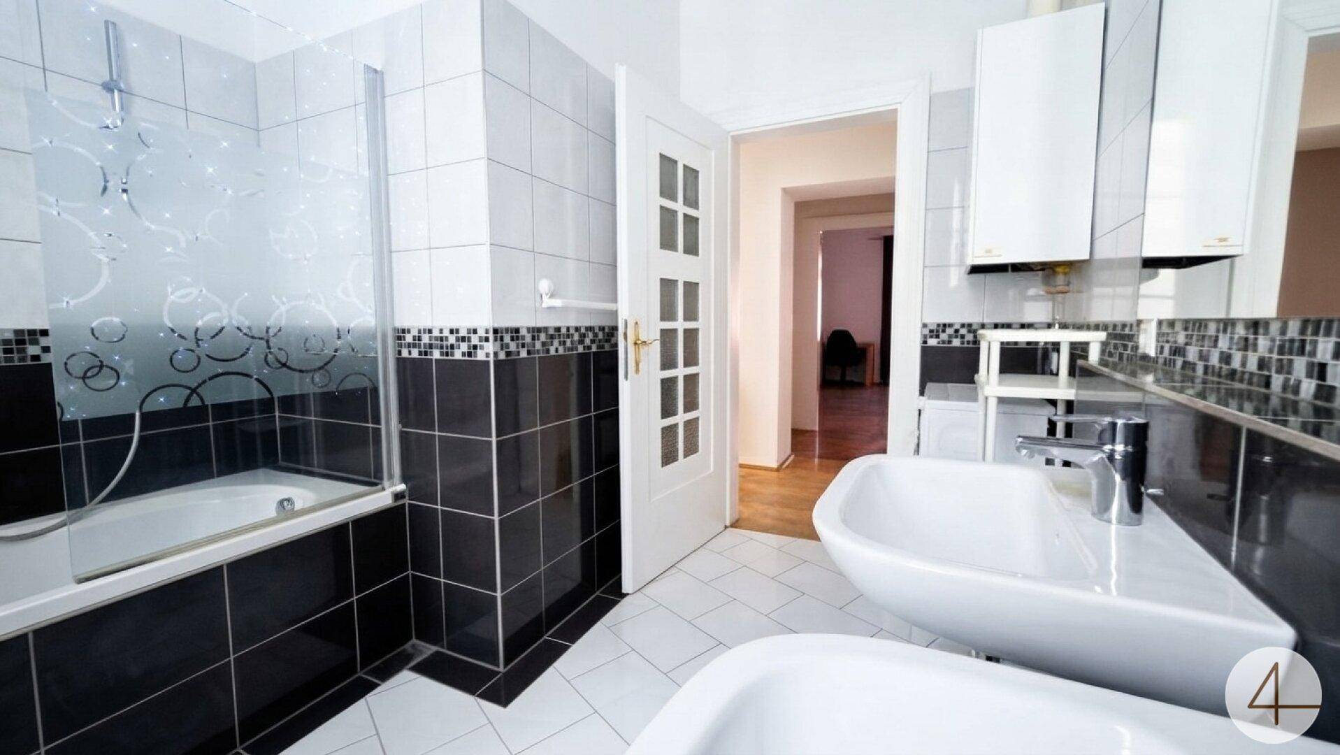 Helles Badezimmer mit Badewanne und Duschabtrennung, Doppelwaschbecken und schwarz-weißen Fliesen.