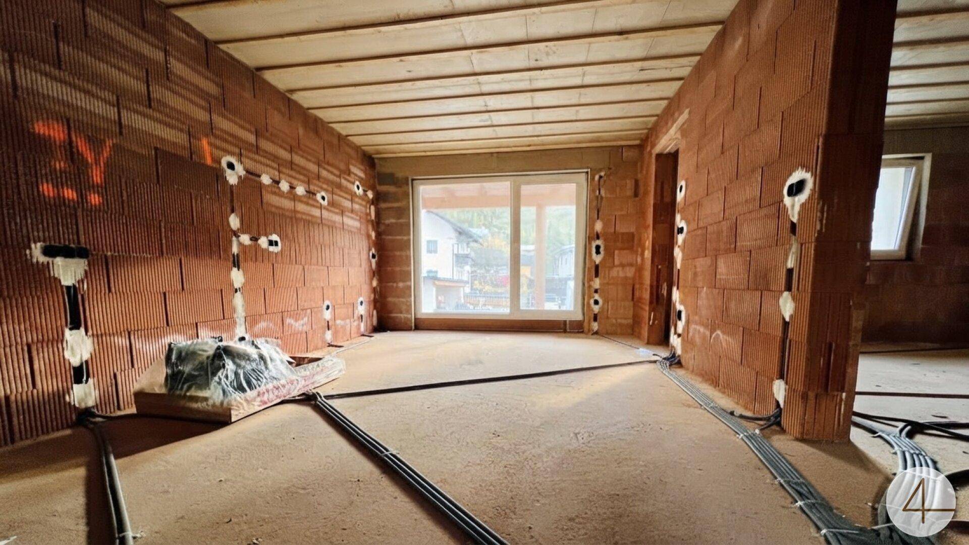 Innenraum im Rohbau mit Ziegelwänden, sichtbaren Leitungen und einem großen Fenster.
