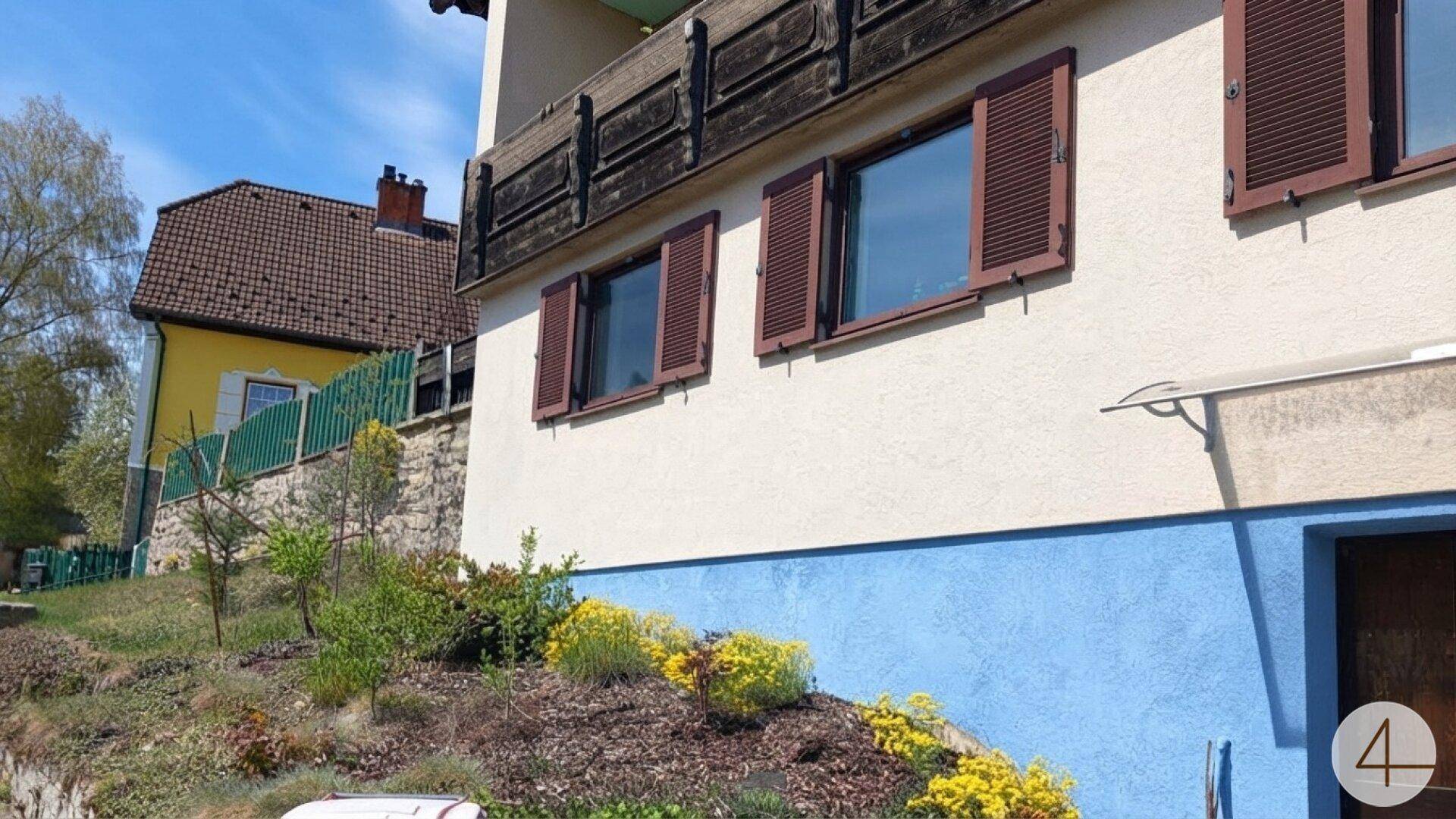 Detailansicht der Hausfassade mit blauer Sockelbemalung, Fensterläden und blühenden Pflanzen im Vorgarten.