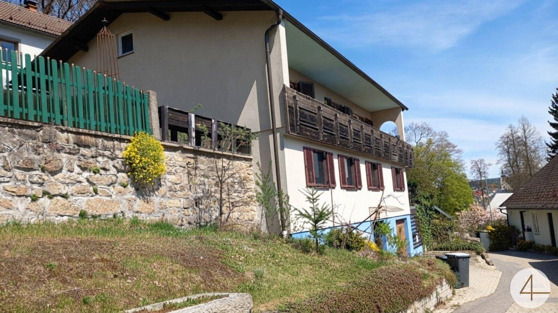 Seitenansicht eines Hauses mit Balkon, Holzgeländer und traditionellen Fensterläden, umgeben von Vegetation.