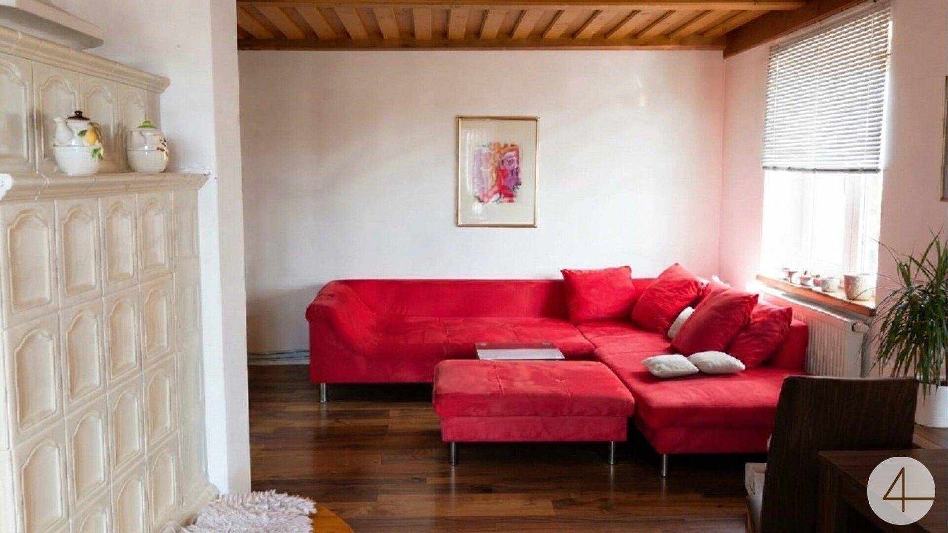 Gemütliches Wohnzimmer mit rotem Ecksofa, traditionellem Kachelofen und Holzbalkendecke.