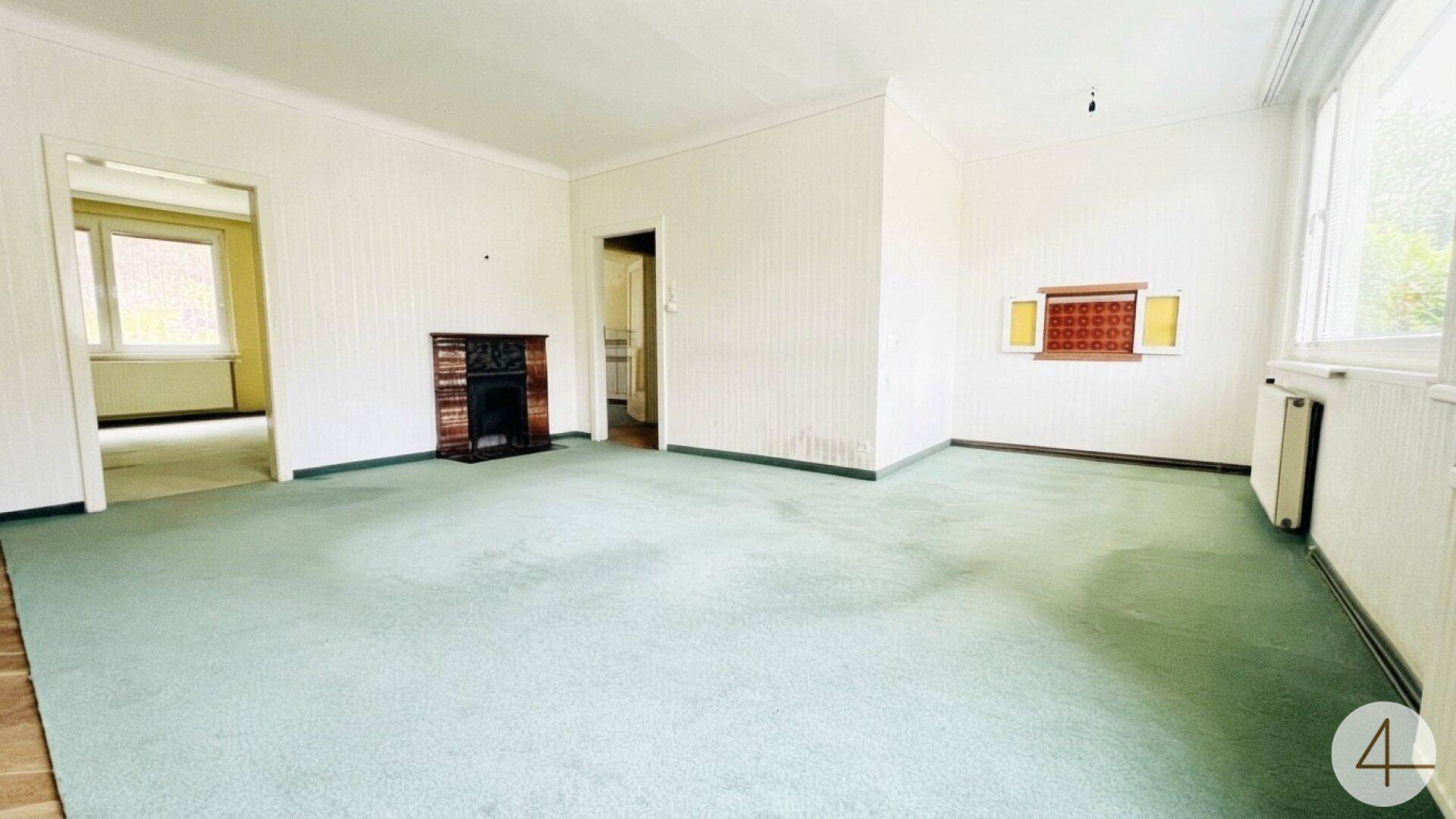 Helles Wohnzimmer mit grünem Teppichboden, Kamin und Zugang zu weiteren Räumen.