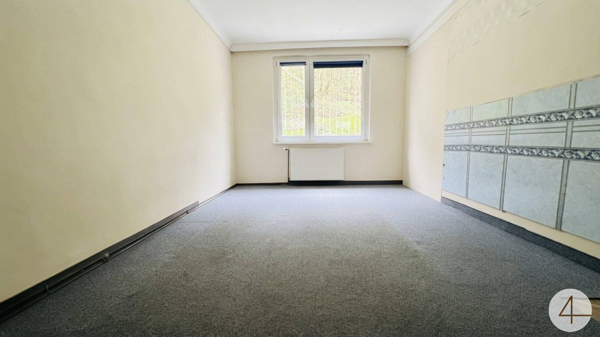 Einfaches Zimmer mit grauem Teppichboden, hellen Wänden und einem Fenster.