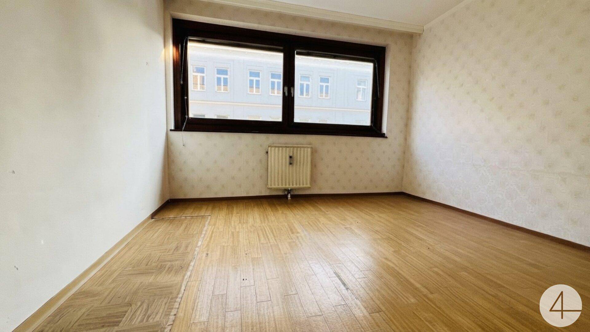 Heller Raum mit Holzparkett, Tapeten und einem Fenster, das Tageslicht hereinlässt.
