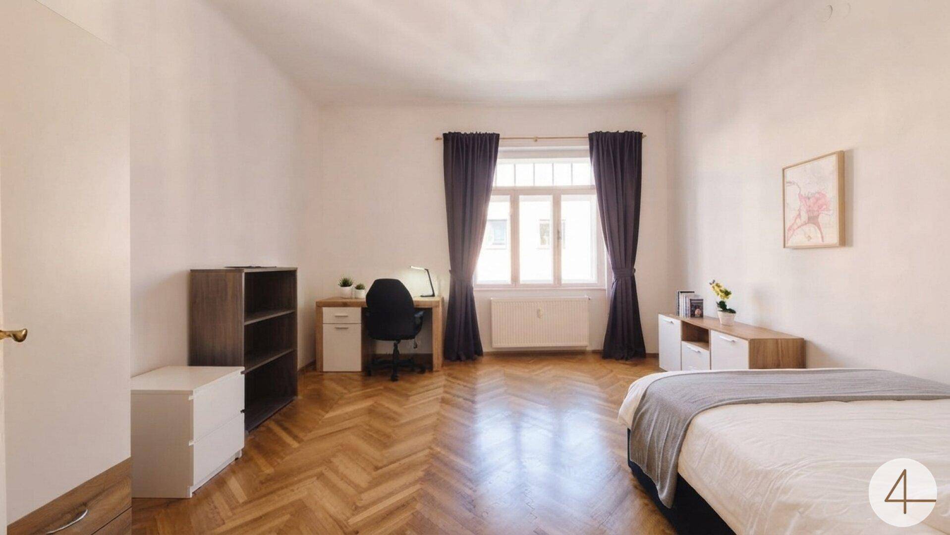 Geräumiges Schlafzimmer mit Schreibtisch, Bett und großem Fenster, ausgestattet mit dunklen Vorhängen.