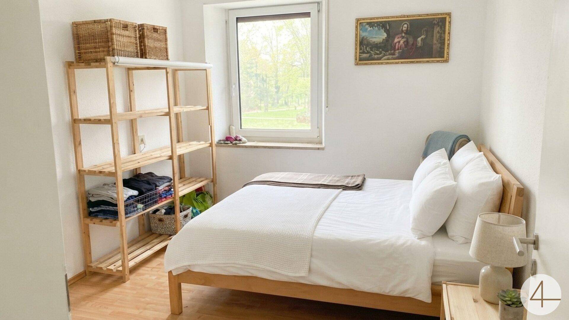 Schlafzimmer mit Holzbett, offenem Regal und einem Fenster mit Blick ins Grüne.