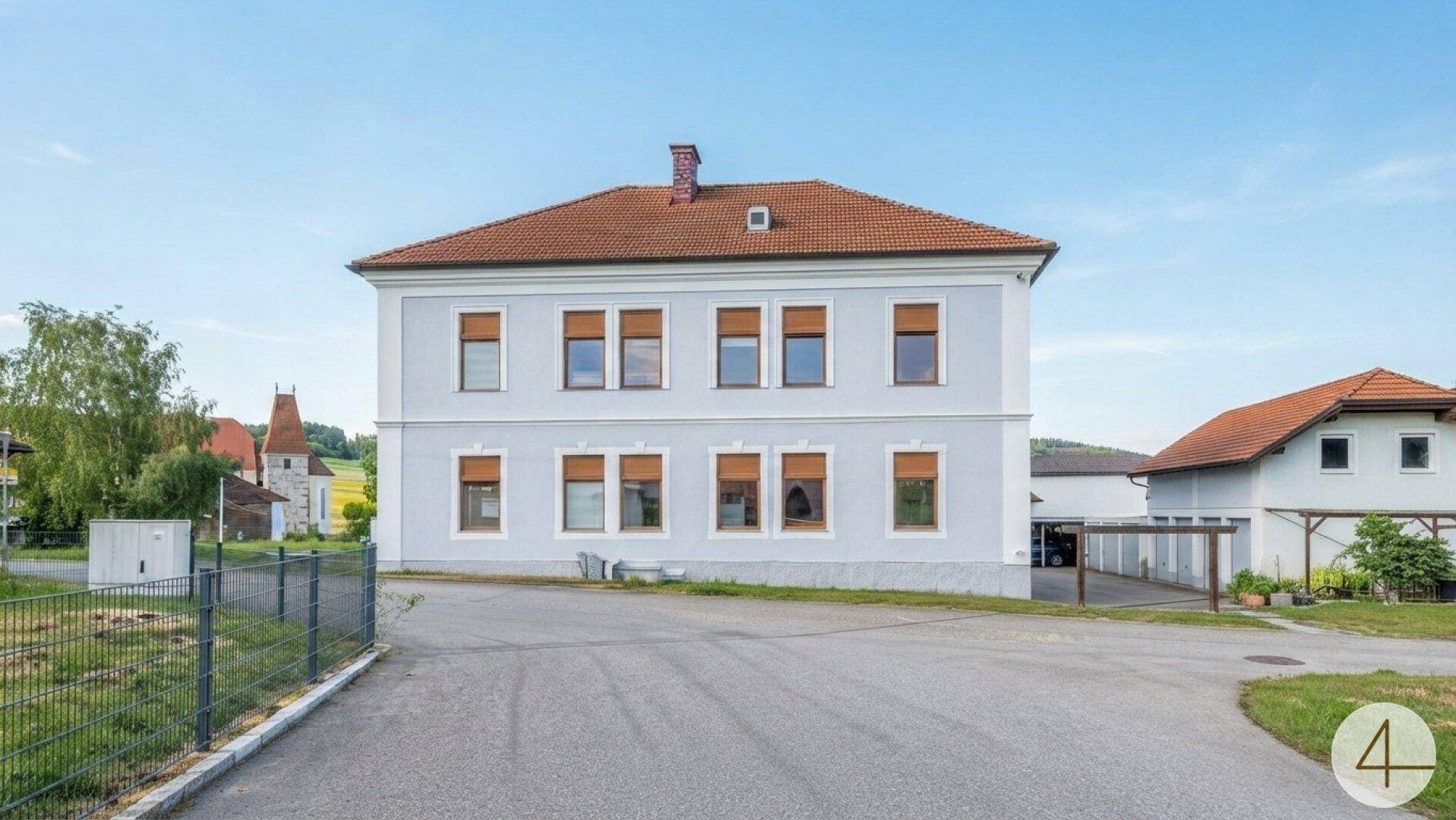 Ansicht eines großen Gebäudes mit hellblauer Fassade, rotem Ziegeldach und einem Vorplatz.
