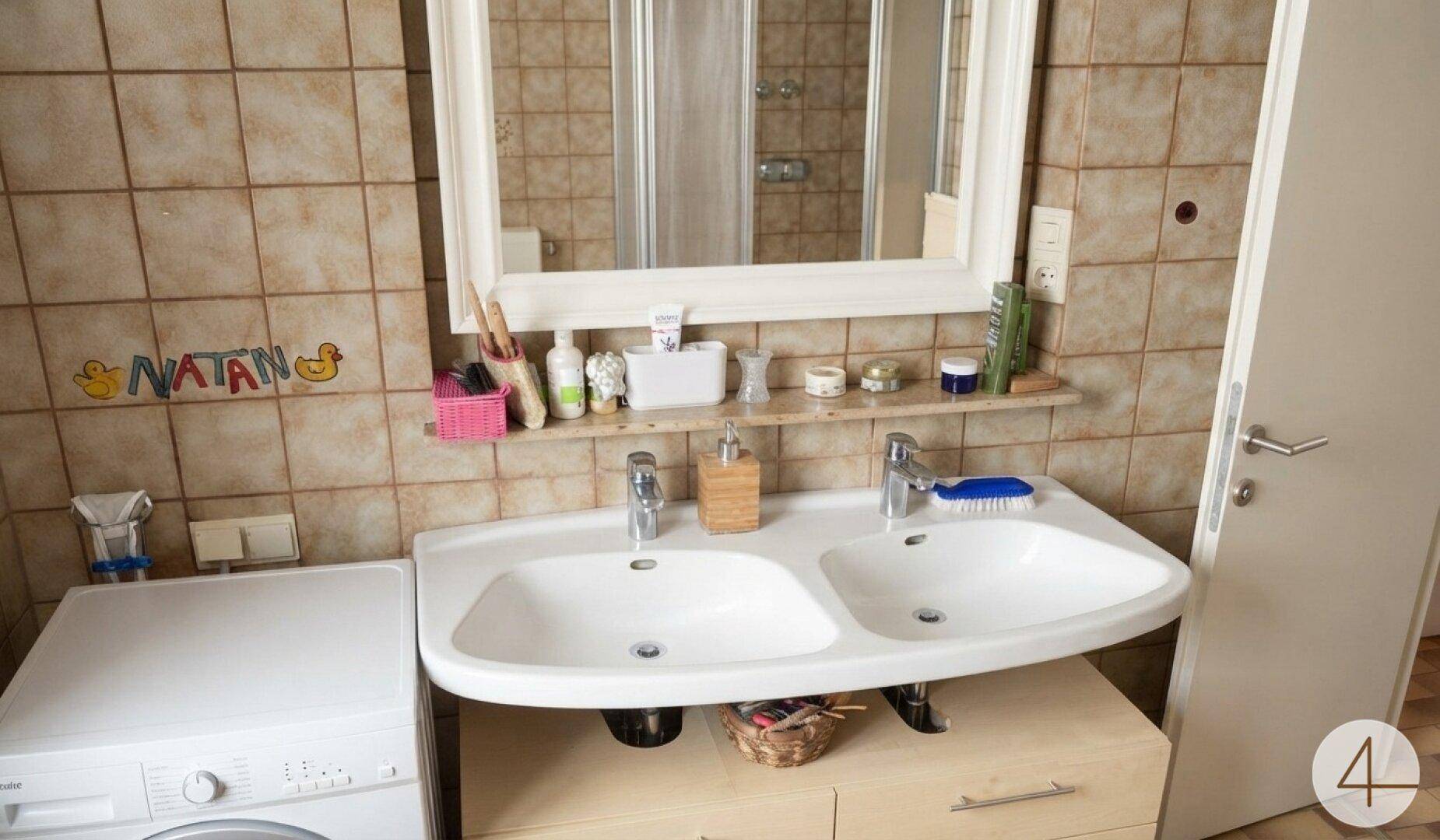 Badezimmer mit Doppelwaschbecken, großem Spiegel und braun gefliesten Wänden, inklusive Waschmaschine.