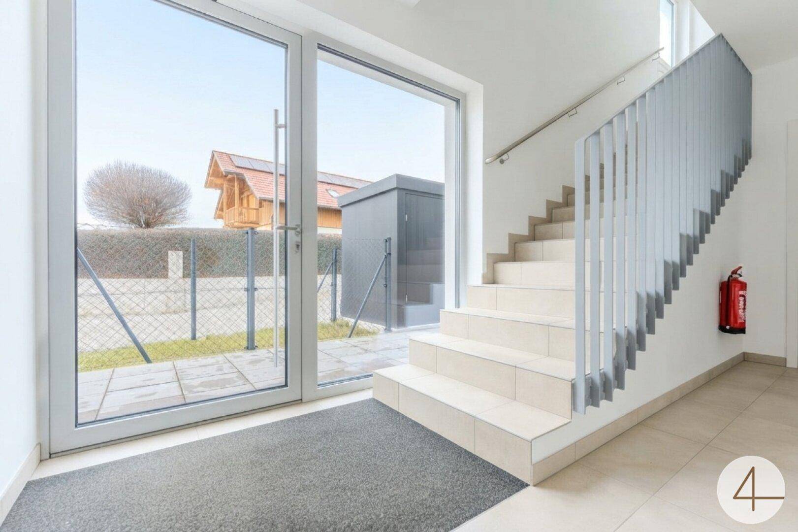 Heller Treppenaufgang in einem modernen Gebäude mit großer Glastür zum Außenbereich.