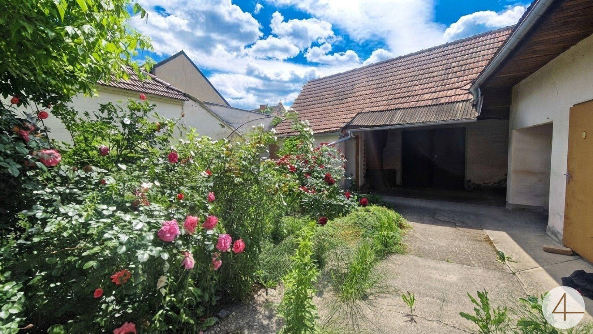 Blühender Rosengarten vor einem älteren Gebäude mit offenem Garagenbereich und gepflastertem Hof.