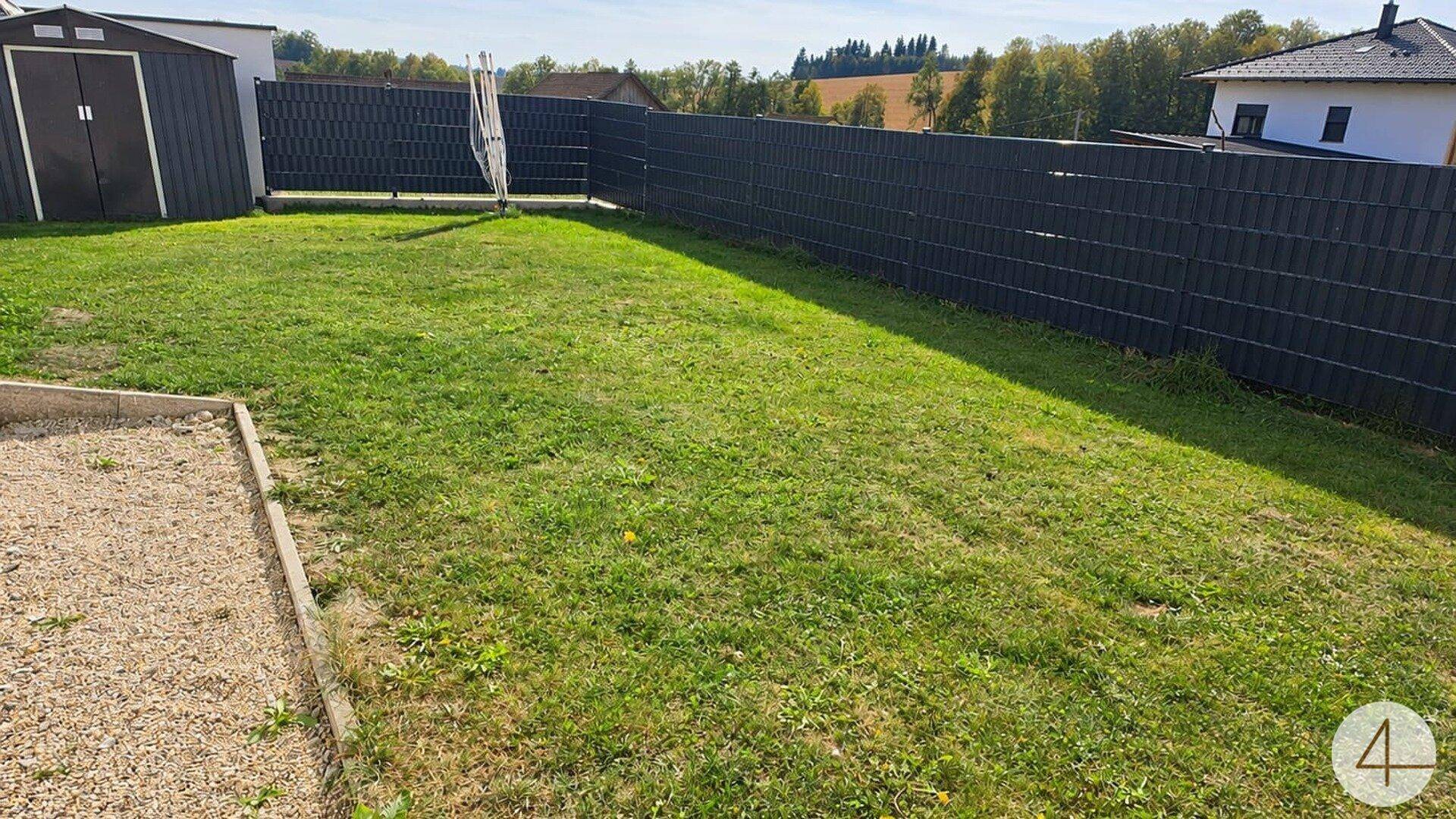 Weitläufiger Gartenbereich mit Rasenfläche, Geräteschuppen und blickdichtem Zaun im Sonnenlicht.