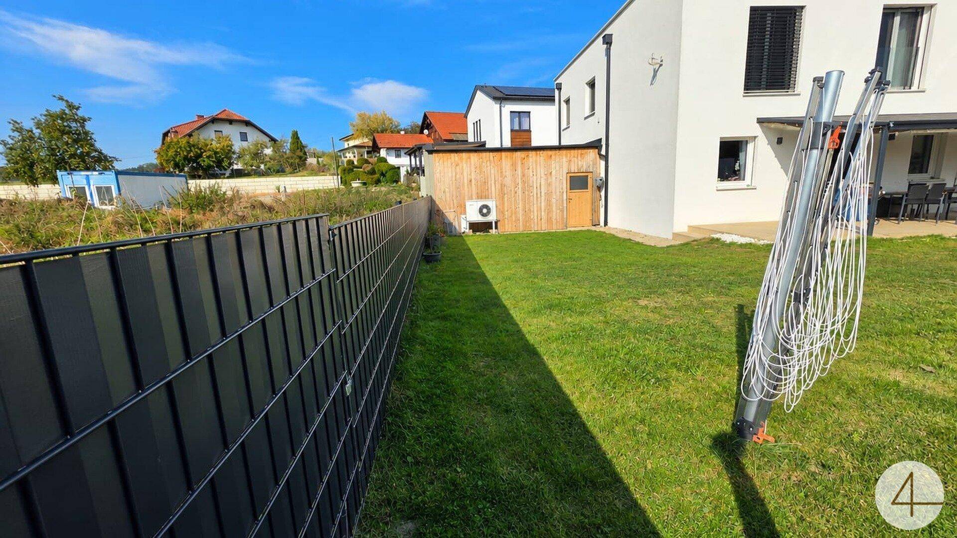 Seitenansicht des modernen Hauses mit Rasenfläche, Zaun und Wäschespinne im sonnigen Garten.
