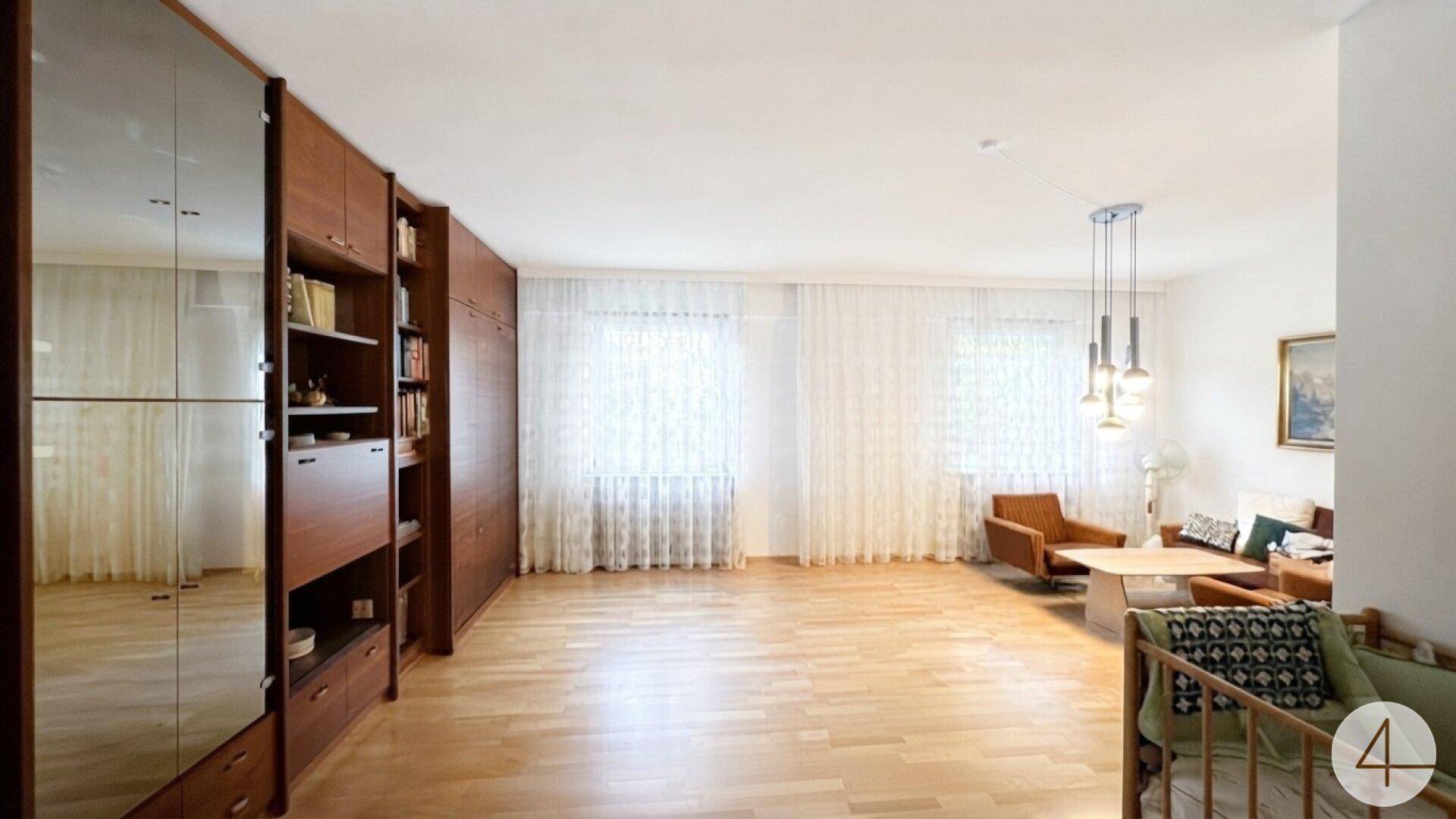 Geräumiges Wohnzimmer mit hellem Parkettboden, großen Fenstern und moderner Beleuchtung.