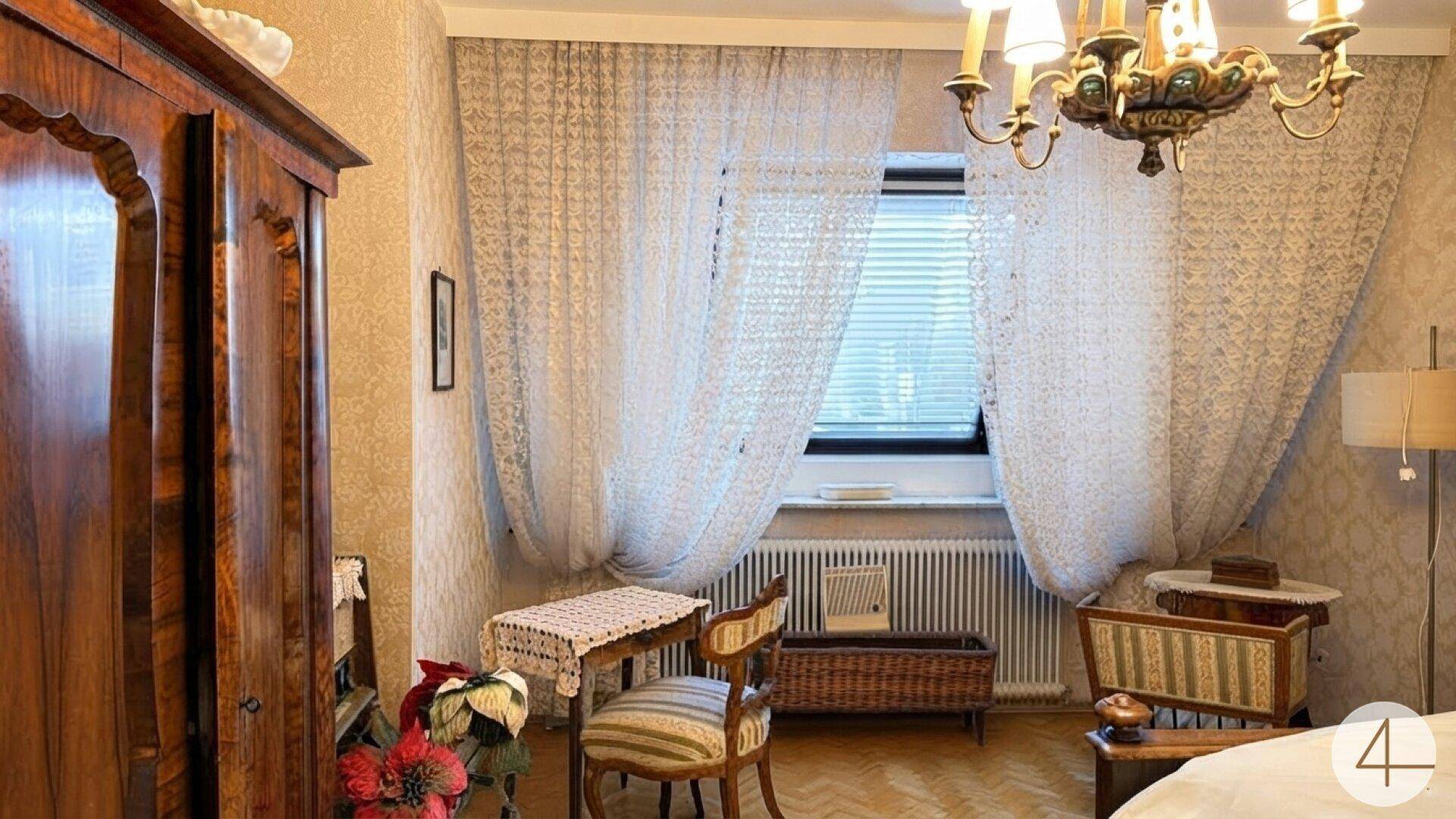 Schlafzimmer mit klassischem Mobiliar, Parkettboden und Fenster mit Vorhängen.