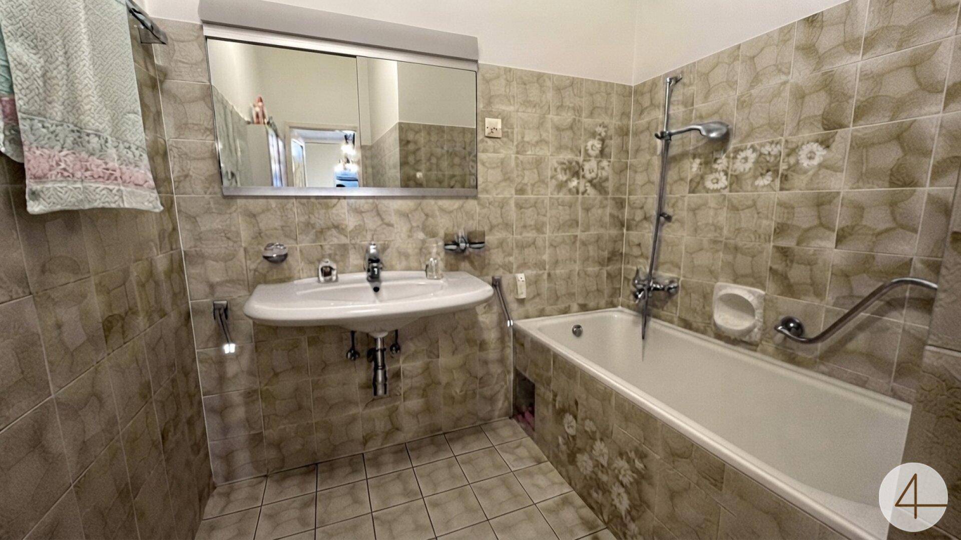 Badezimmer mit Badewanne, Waschbecken und gemusterten Fliesen im 80er-Jahre-Stil.