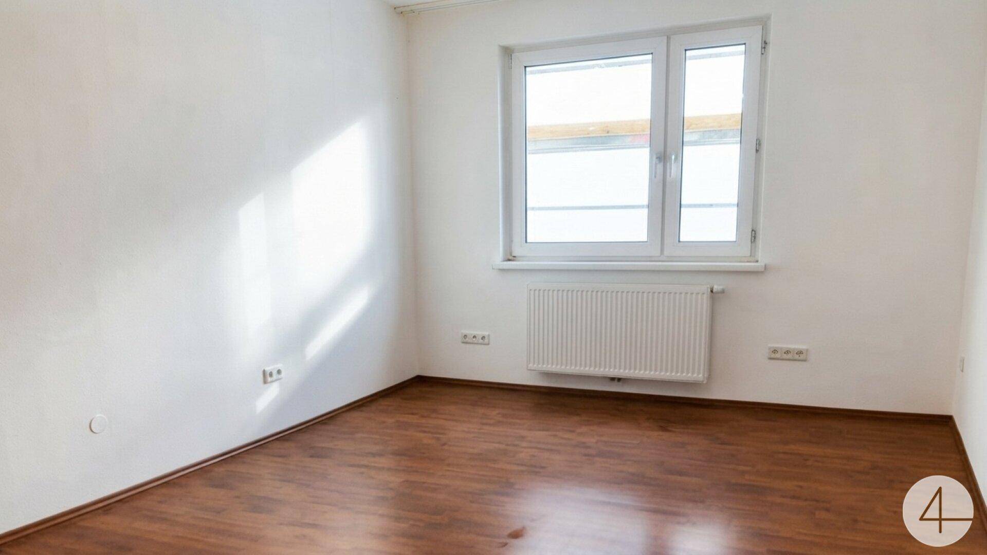 Leeres Zimmer mit Laminatboden, weiß gestrichenen Wänden und einem großen Fenster.