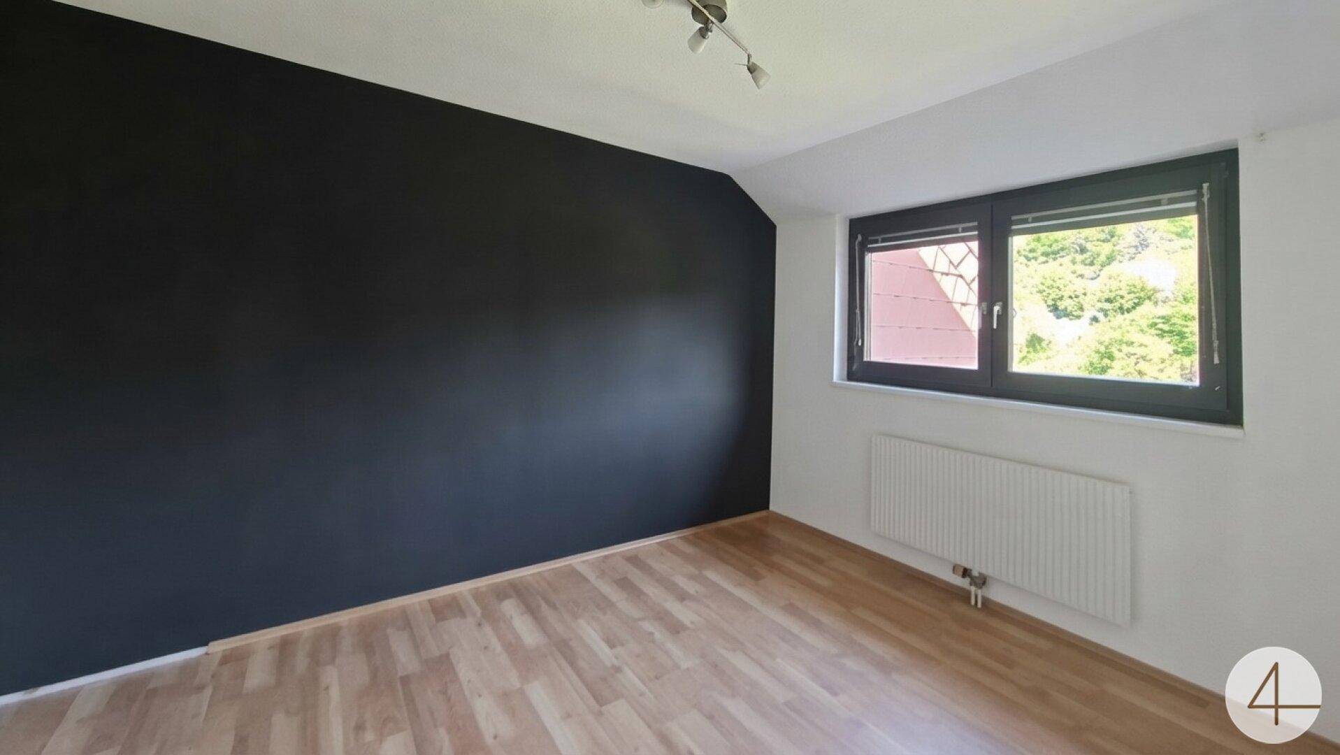 Leeres Zimmer mit dunkler Akzentwand, Holzfußboden und Fenster für natürliches Licht.
