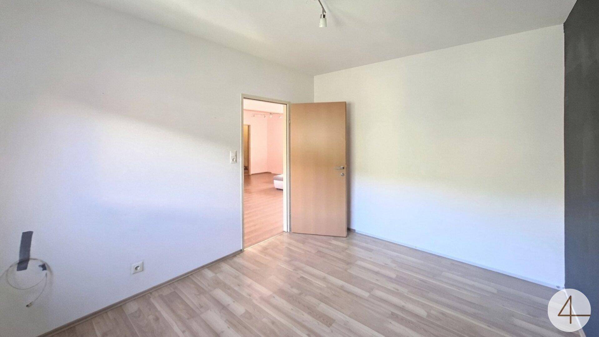Helles, leeres Zimmer mit Holzfußboden und Zugang zu einem angrenzenden Wohnbereich.
