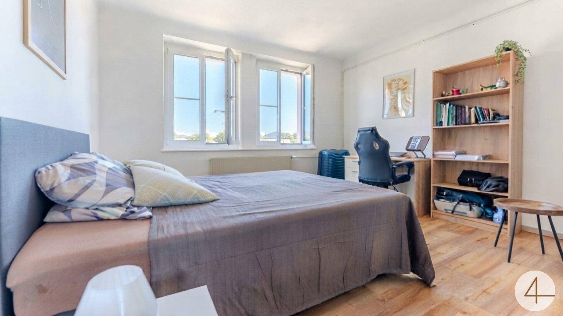 Schlafzimmer mit Bett, Schreibtisch, Bücherregal und zwei großen Fenstern.