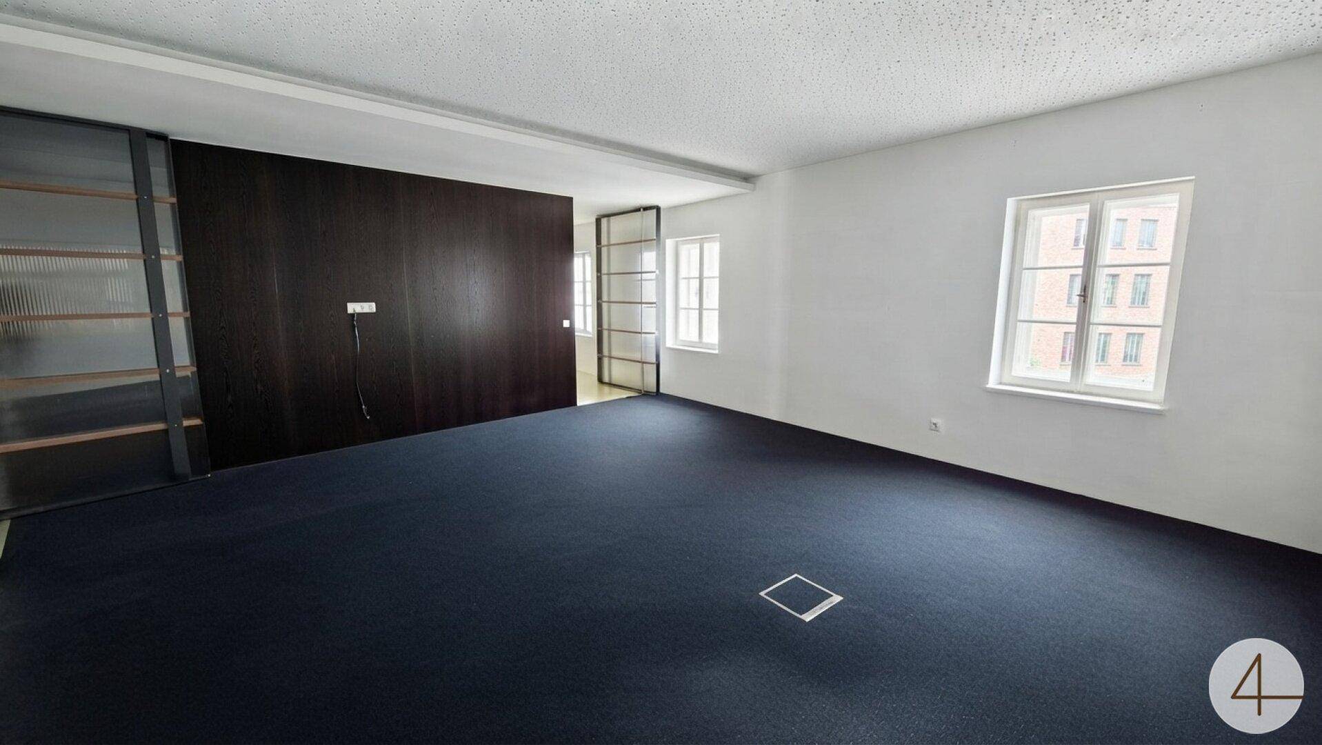 Großer Büroraum mit dunklem Teppichboden, Holzwand und Fenstern mit Blick nach draußen.
