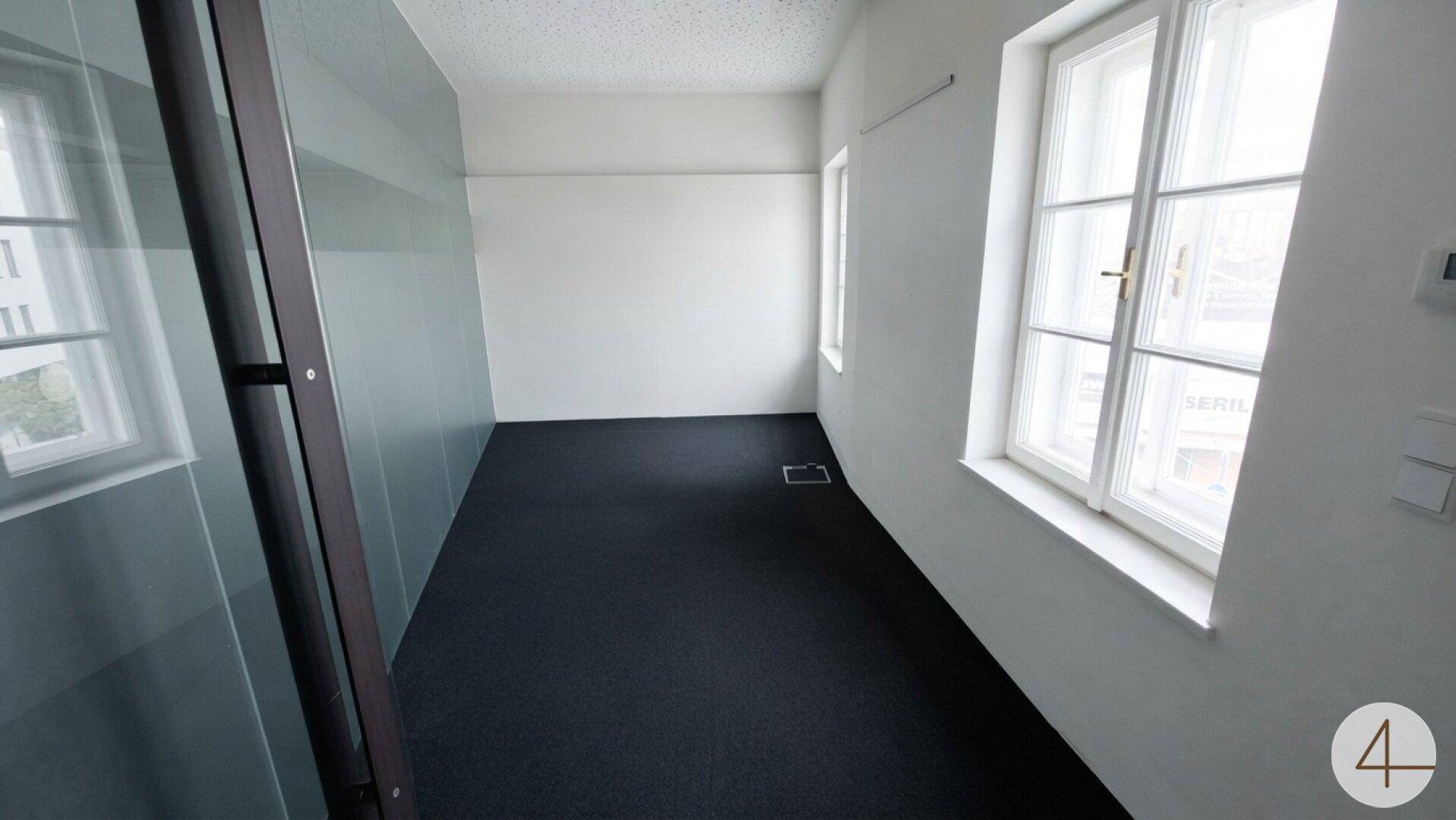 Schmaler Büroraum mit Teppichboden, großen Fenstern und einer Glaswand.