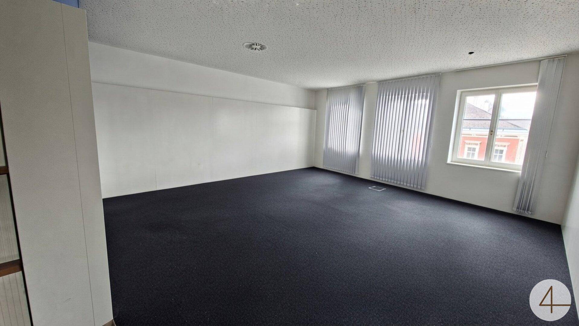 Heller Büroraum mit Teppichboden und vertikalen Jalousien an den Fenstern.