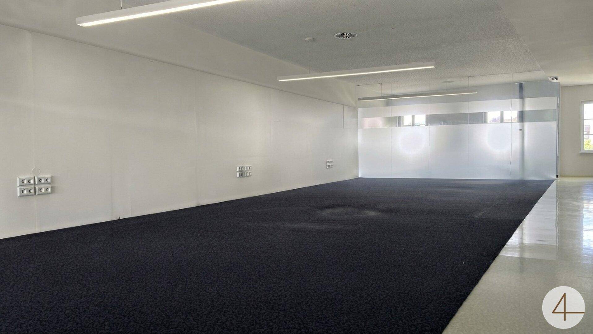 Großer, offener Bürobereich mit dunklem Teppichboden und langen Leuchtstoffröhren an der Decke.