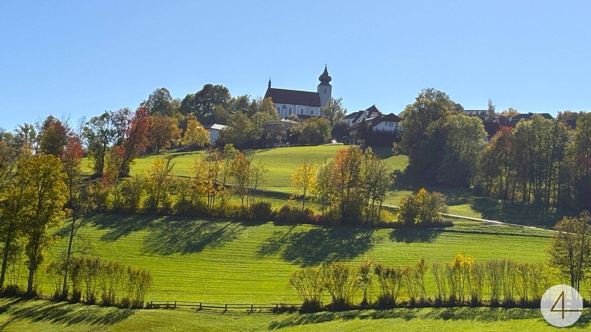 Malerische Landschaft mit einem Dorf und einer Kirche auf einem Hügel, umgeben von grünen Wiesen.