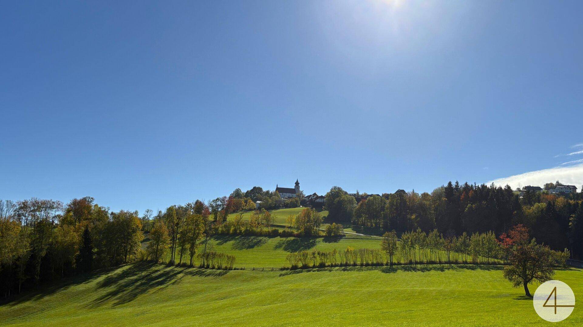 Weitläufige Panoramalandschaft mit einem Dorf und einer Kirche auf einem sonnigen Hügel.