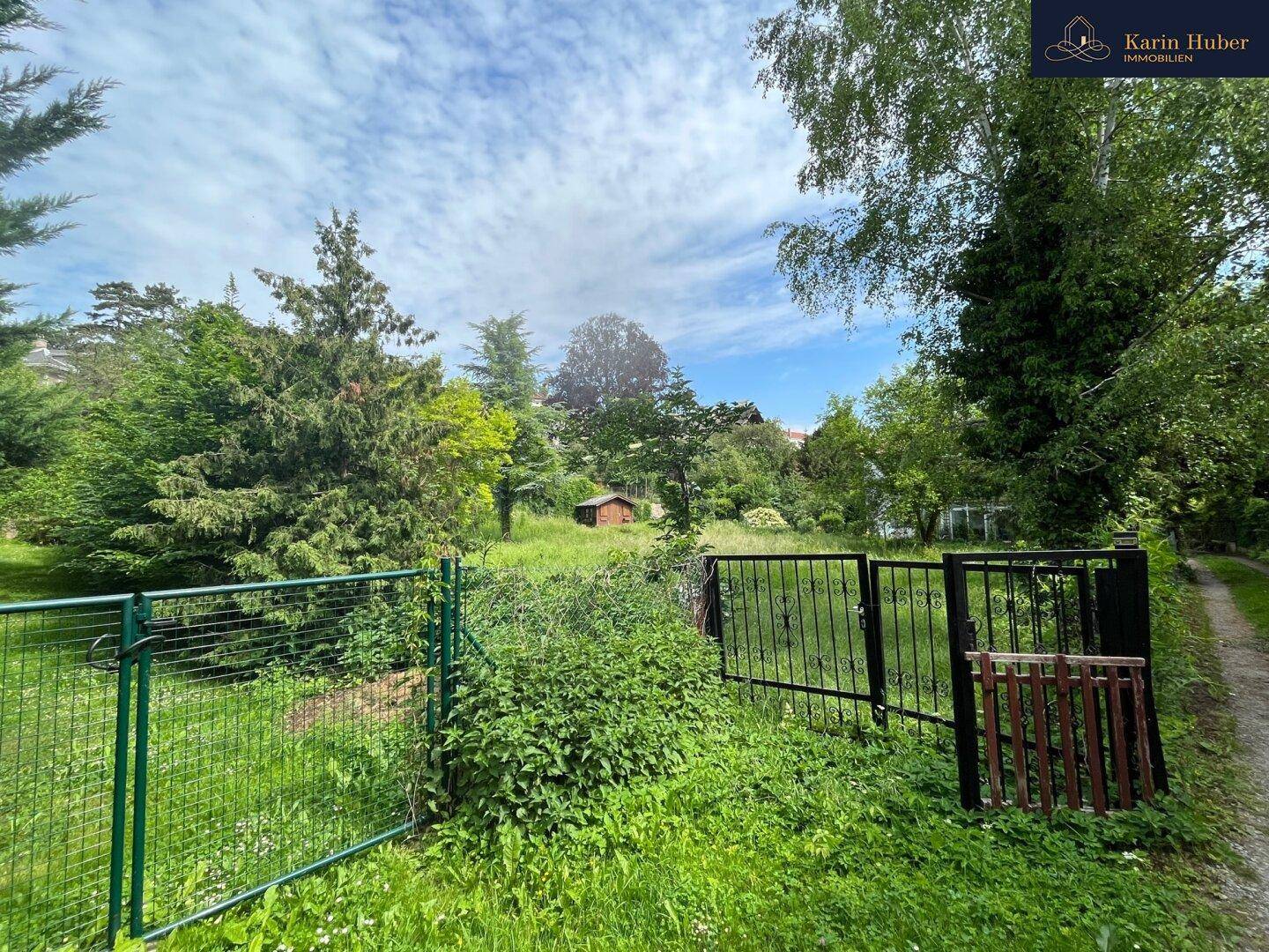 Großes Gartengrundstück mit Zaun und Tor, umgeben von Bäumen und Sträuchern unter bewölktem Himmel.