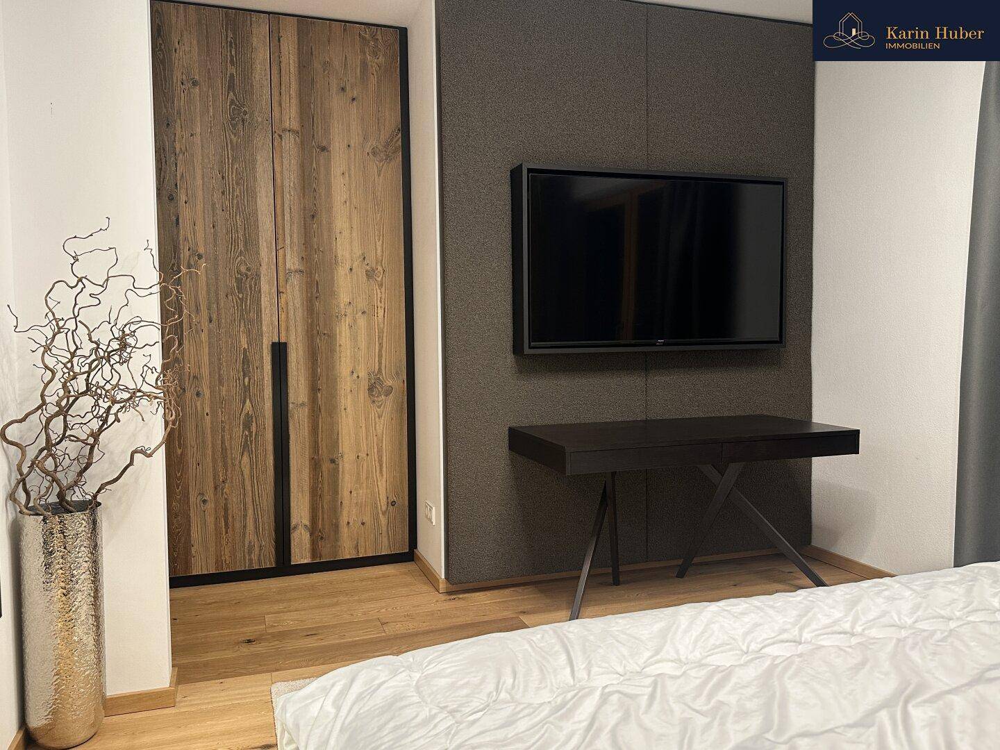 Schlafzimmer mit Holzkleiderschrank, Wandfernseher und modernem Schreibtisch.