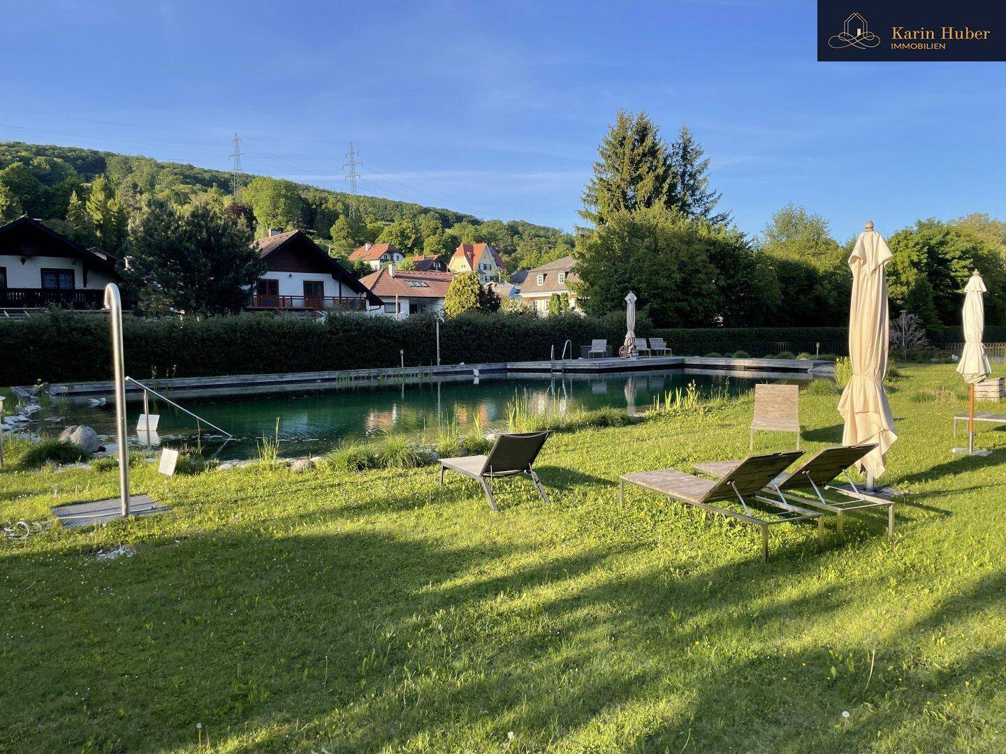 Natürlicher Schwimmteichbereich mit Liegestühlen und Sonnenschirmen, umgeben von Grünflächen.
