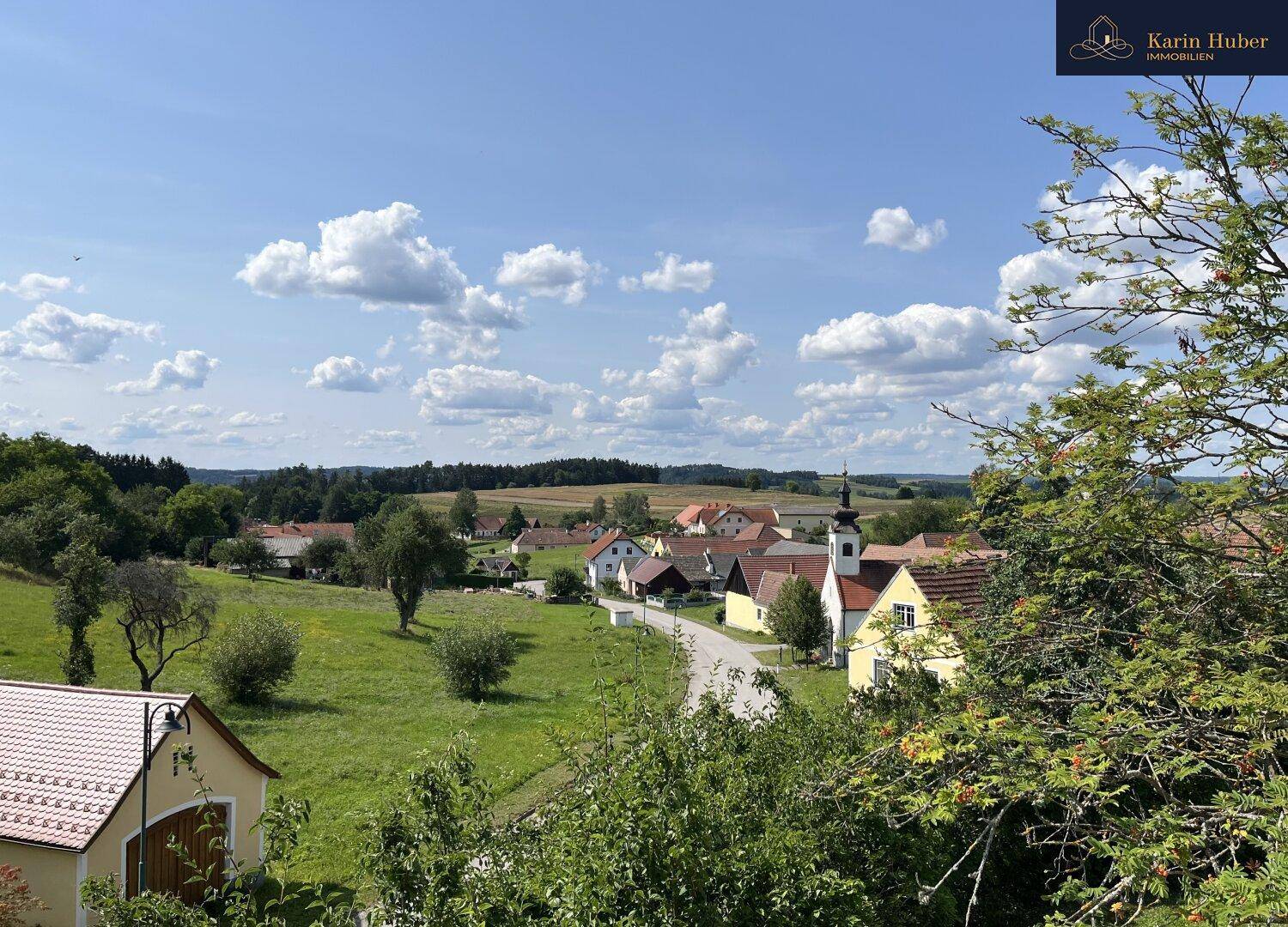 Malerische Landschaft mit weiten grünen Feldern, kleinen Häusern und einem Kirchturm unter blauem Himmel.