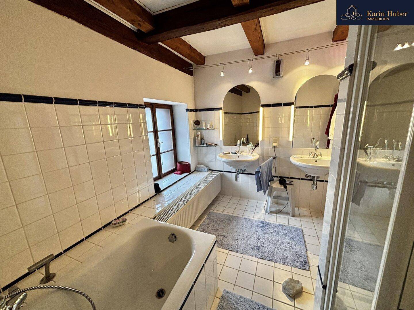Badezimmer mit weißen Fliesen, Badewanne, zwei Waschbecken und einem Fenster.