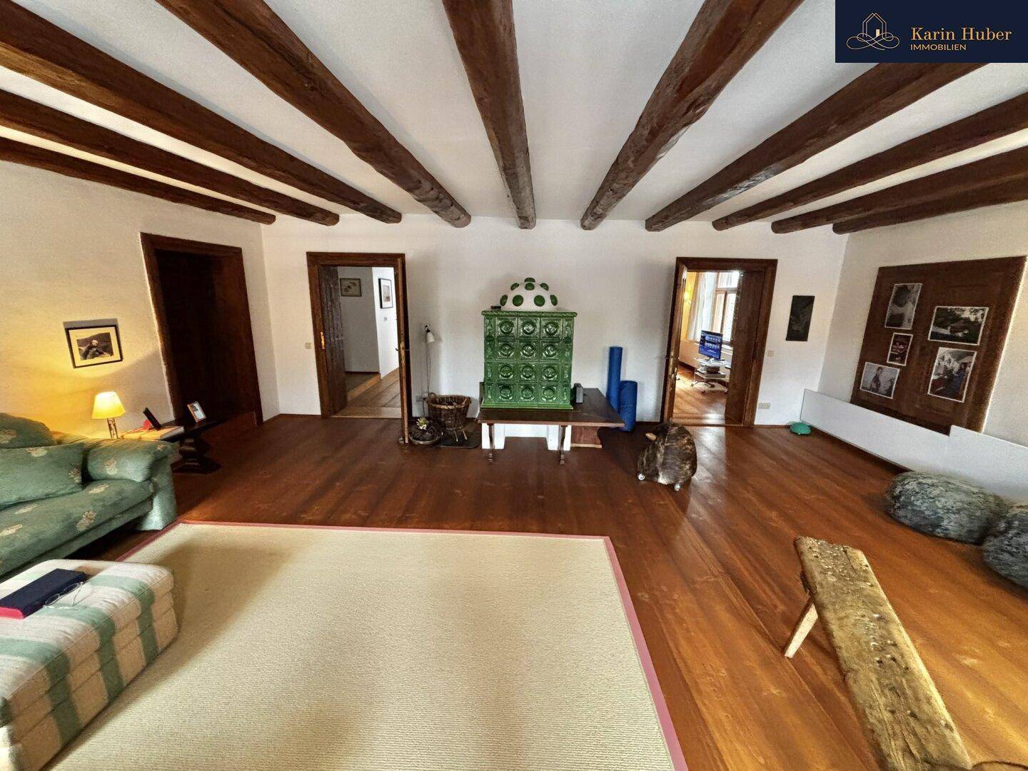Großes Wohnzimmer mit Holzbalkendecke, Holzboden und einem grünen Kachelofen als Blickfang.