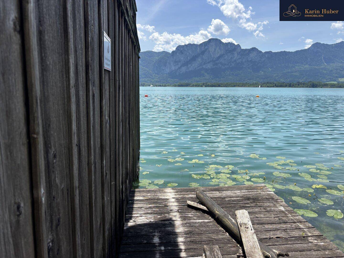 Seitenansicht eines Holzbootshauses direkt am Wasser mit Seerosen und Bergpanorama im Hintergrund.