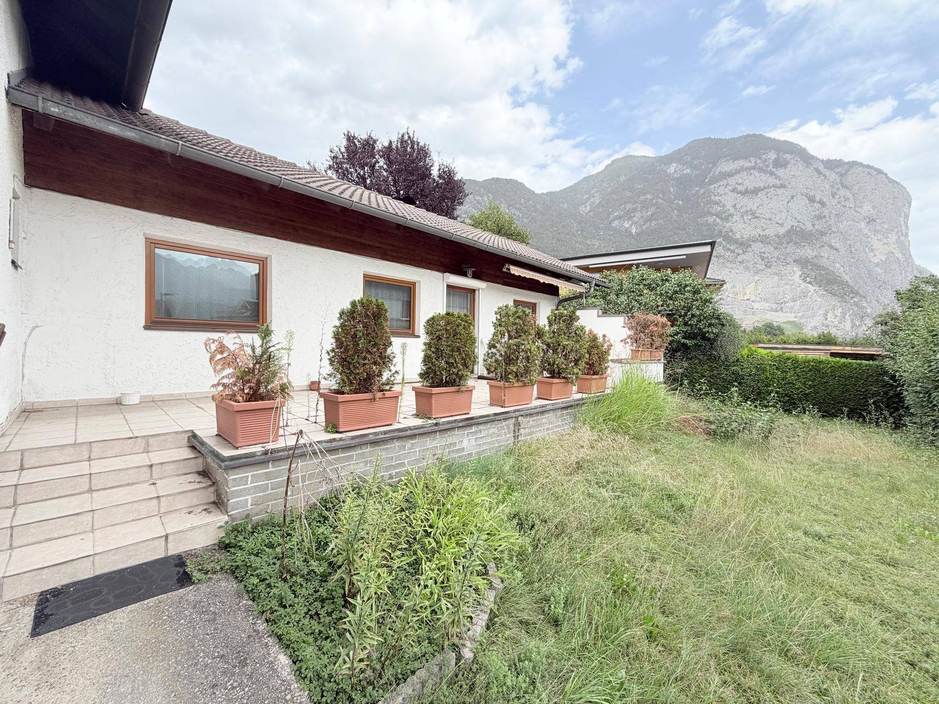 Terrasse am Haus mit Topfpflanzen und Blick auf den verwilderten Garten und die Berge unter bewölktem Himmel.