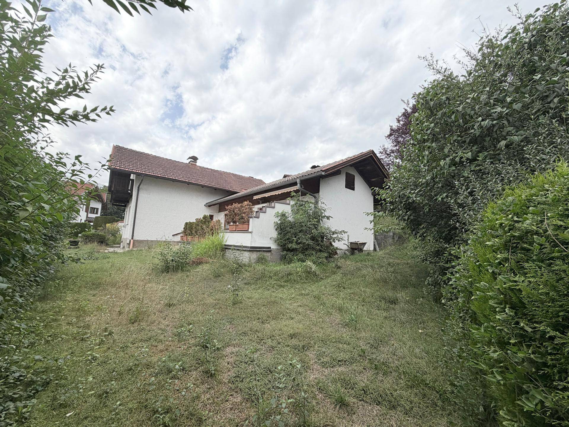 Hinteransicht des Hauses auf einem grünen Hanggrundstück mit üppiger Vegetation unter bewölktem Himmel.