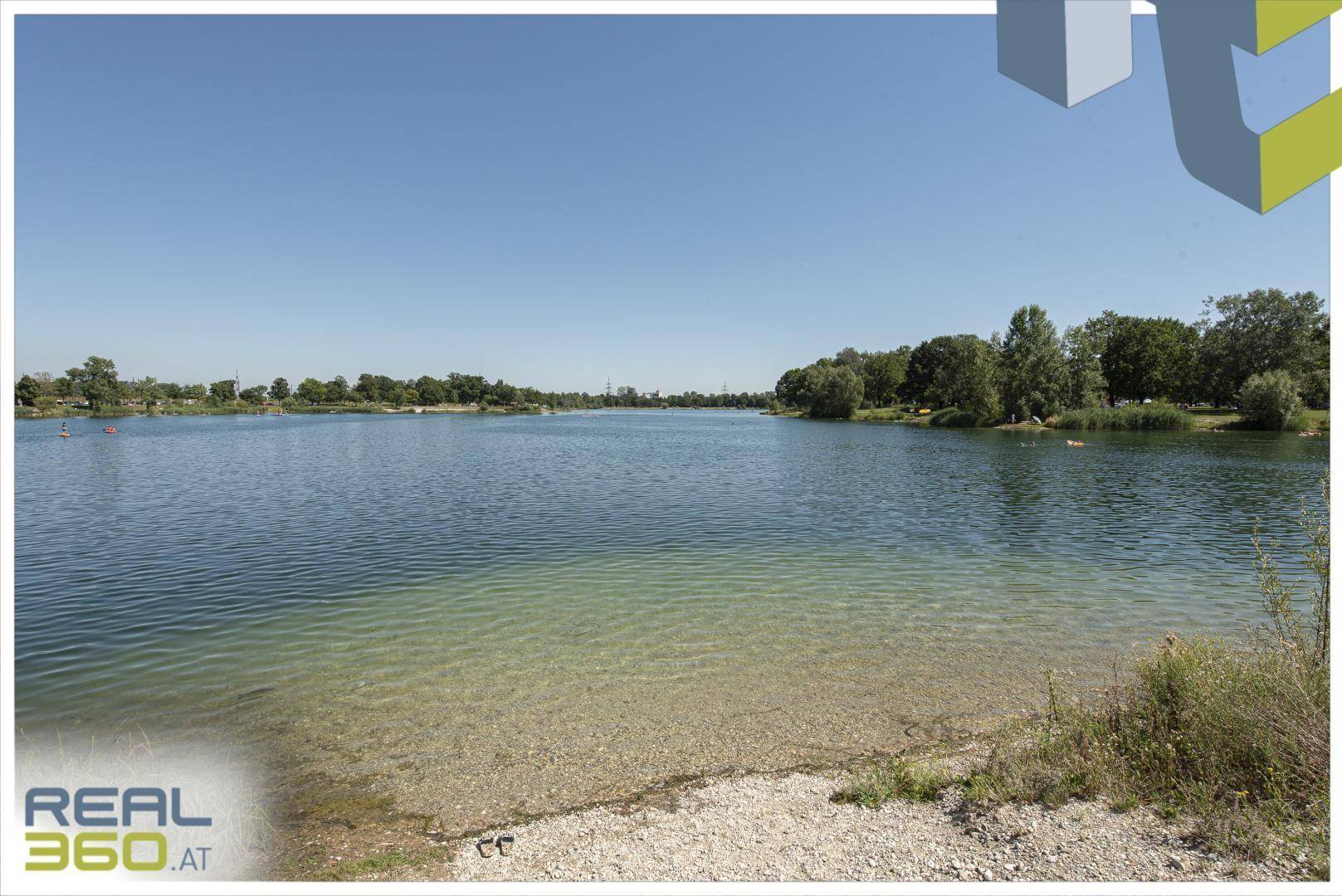 Klarer Badesee mit sandigem Ufer und grünen Bäumen unter einem weiten blauen Himmel.