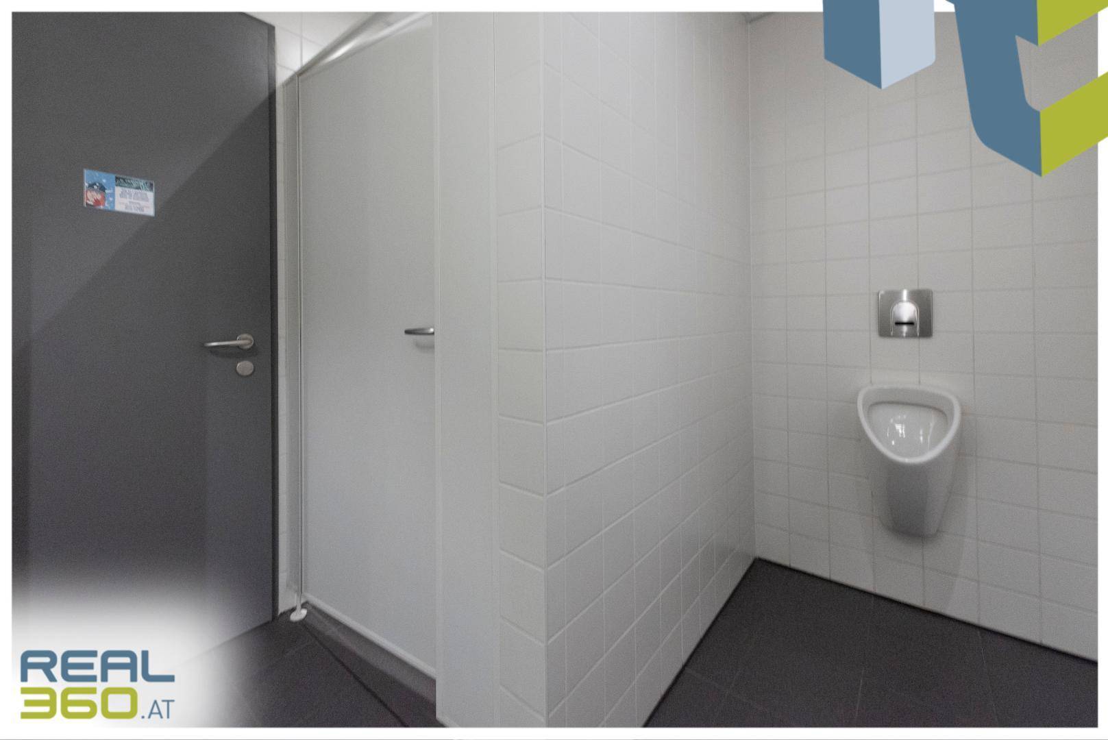 Toilettenbereich mit Urinal, weißen Fliesen und einer grauen Tür, sauber und funktional.