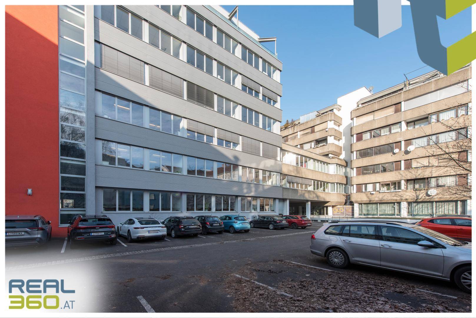 Modernes Bürogebäude mit vielen Fenstern und einem Parkplatz davor unter blauem Himmel.