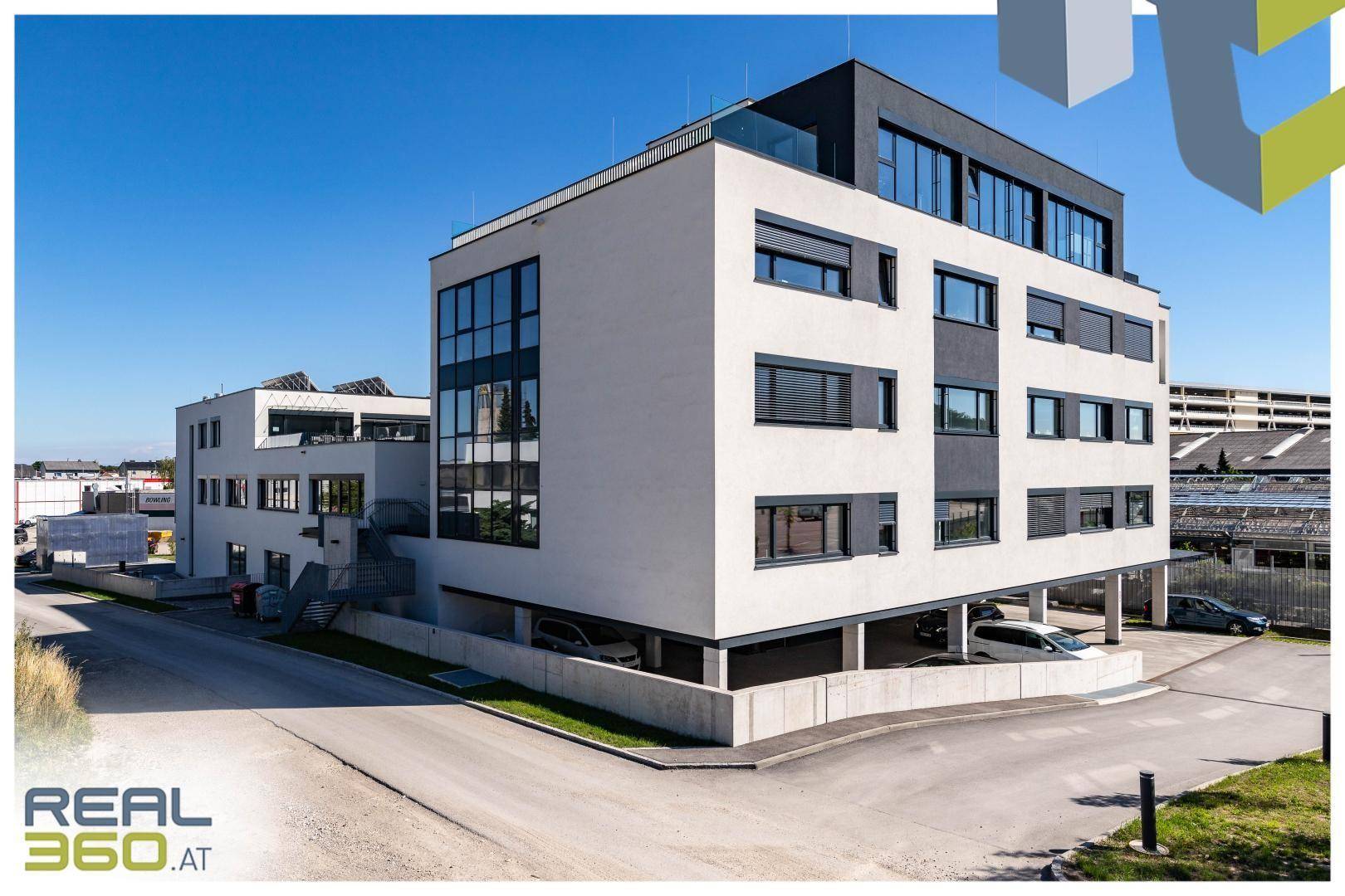Seitenansicht eines modernen Bürogebäudes mit dunklen Fensterrahmen und überdachten Parkplätzen im Freien.